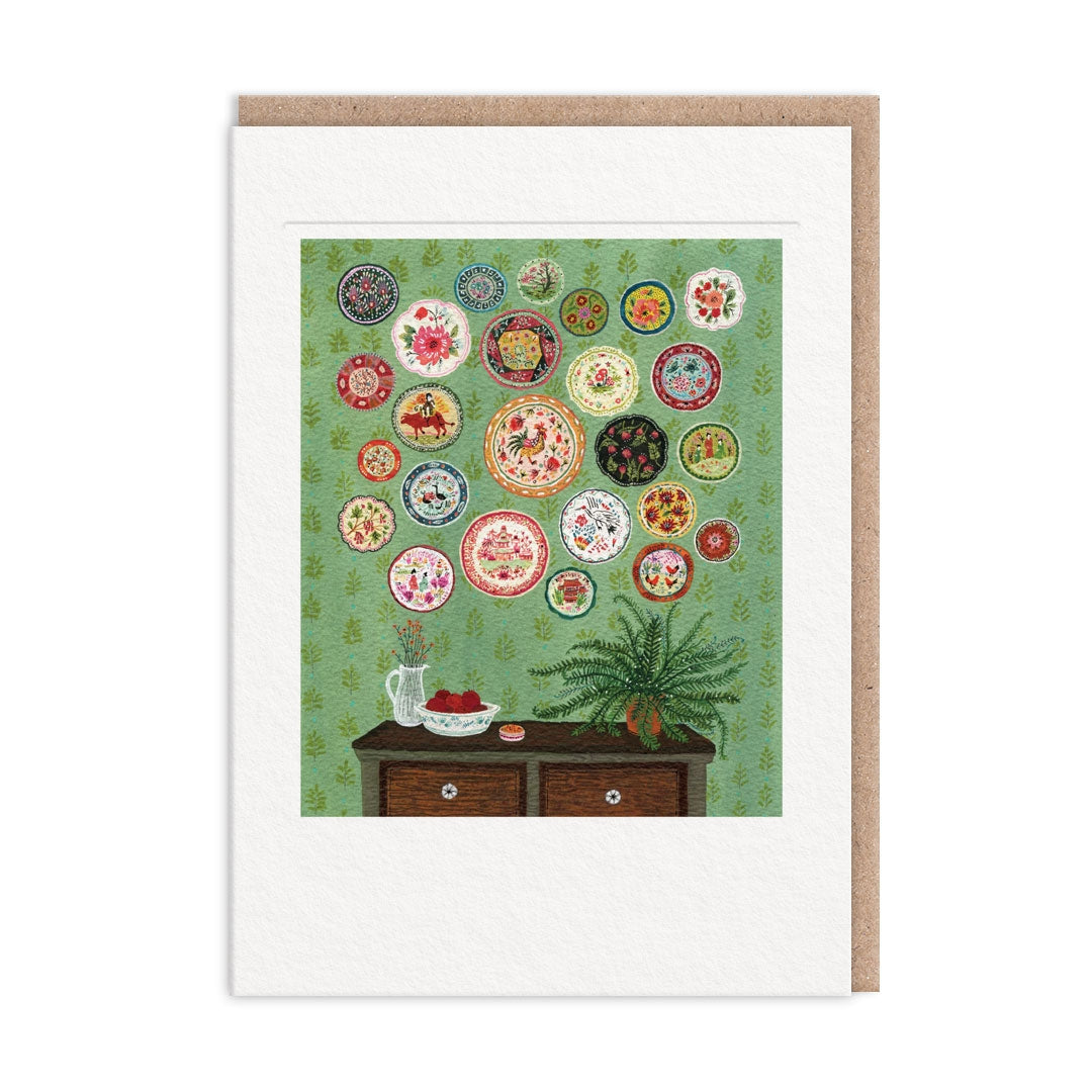 Greeting card showing artwork of a wall with a decorative plate hanging arrangement and a table on a green background. The image is surrounded by a white border.