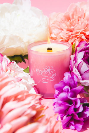 White candle in a pink glass container with purple floral design surrounded by pink and purple flowers on a pink background.