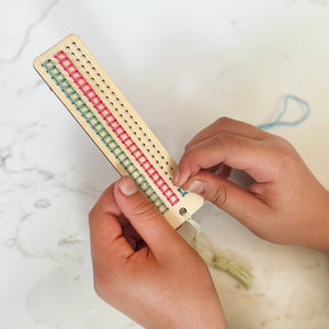 Person adding stitching to a wooden bookmark with colorful threads on a marble surface.