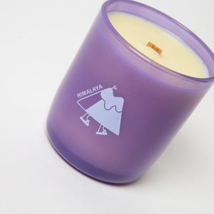White candle in a purple glass container with the text 'Himalaya' and an abstract illustration of a mountain with legs. The candle is on a white background.