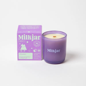 Milkjar candle in a purple container placed beside purple packaging with green branding on a white background.