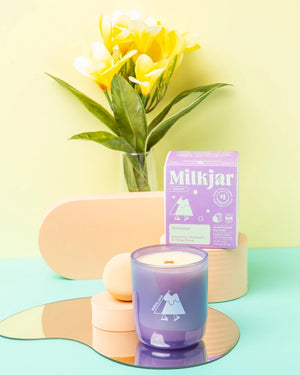 White candle in a purple glass container in front of a purple box showing green Milkjar branding. Both items are on a light blue surface with a vase of flowers in the background. 