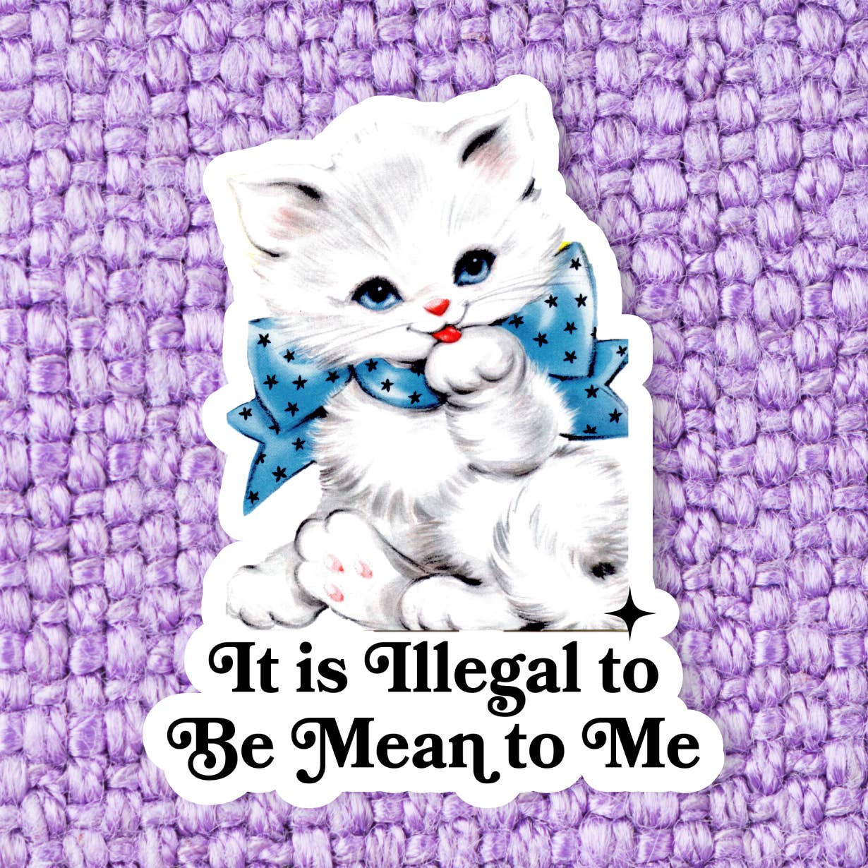 A sticker with an illustration of a white and grey cat wearing a blue, star patterned bow around it's neck as it licks it's paw. The text 'It is illegal to be mean to me' is printed under the cat in black, on a white background. The sticker itself sits on a purple background. 