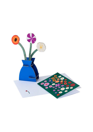 3D pop-up card in the shape of a blue vase containing three colourful flowers. The vase sits alongside a floral greeting card on a white background. 