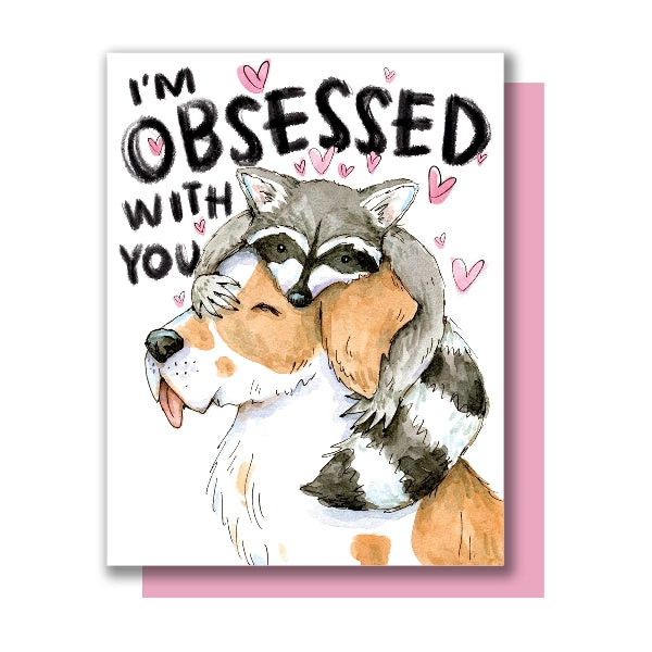 Greeting card with an illustration of a raccoon sitting on a dog's head with the text 'I'm obsessed with you'. The image and text are on a white background with some pink hearts. 