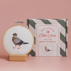 Cross-stitch kit with a completed design of a pigeon in a wooden hoop, next to its packaging on a pink background.