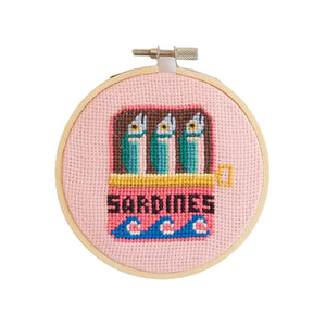 Cross-stitched design of sardines in a tin on a pink background, framed in a wooden hoop.