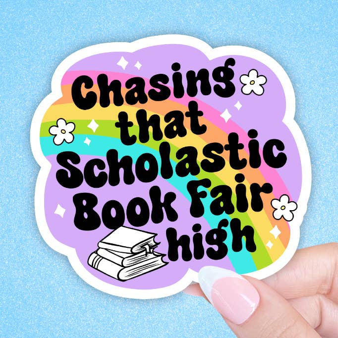 A sticker with rainbow coloured stripes inside a white border. The text 'Chasing that Scholastic book fair high' is printed in black across the sticker. Illustratons of flowers and a stack of books are placed around the text. The sticker is held against a blue background. 