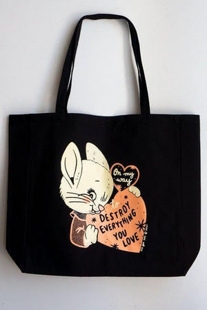 large black tote bag with an orange, black, and off-white design. a smiling, mischievious white rabbit with a sweater holds a heart. another heart is connected to the bunny's head and the large heart. texts in the hearts read "on my way to destroy everything you love"