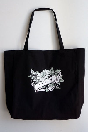 large black tote bag with a white design. a flower, leaves, and a strawberry peak around a banner with the text "yikes"